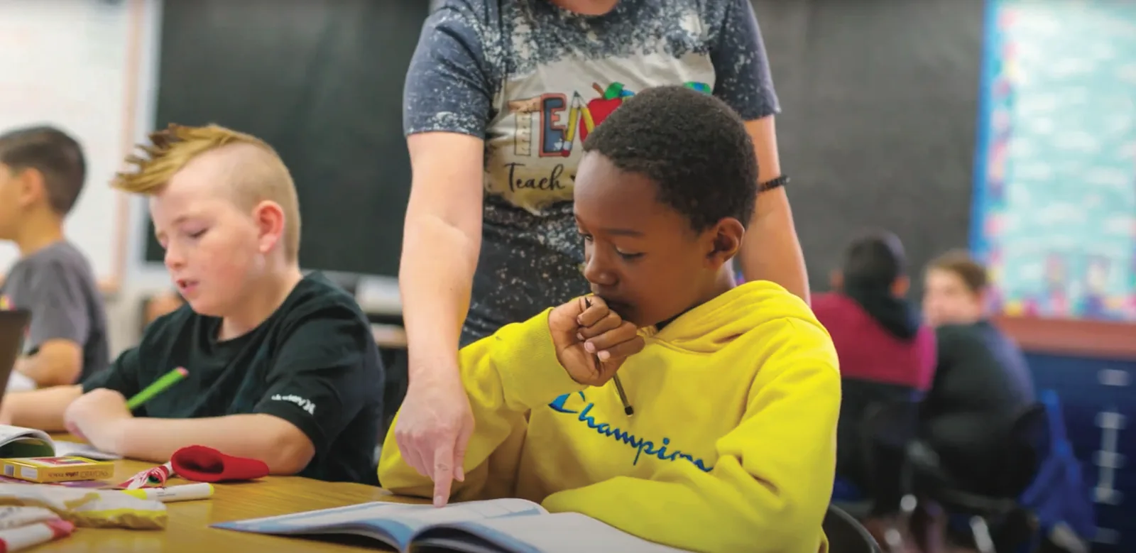 A teacher points into an open book while a student reads along